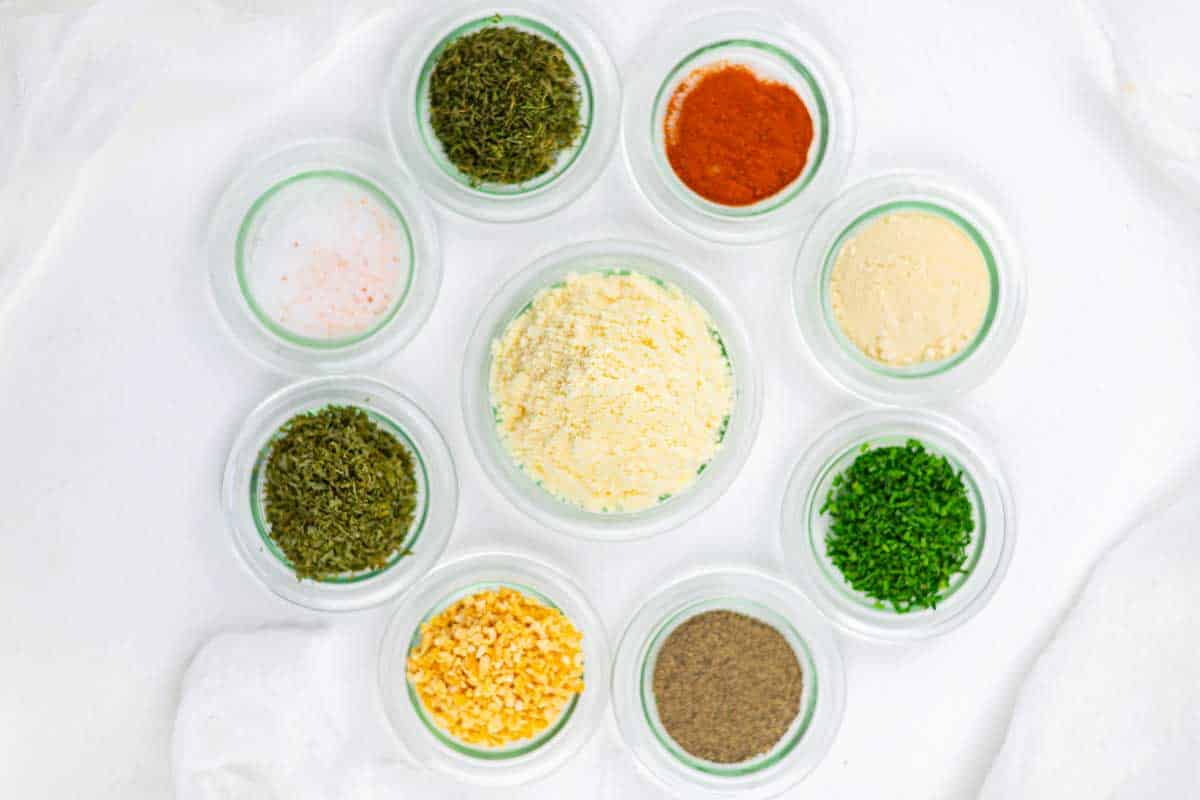 Nine small glass bowls, each containing a different dried herb or spice, arranged in a circle on a white surface.