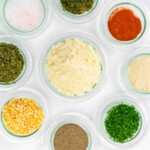 Nine small glass bowls containing various dried herbs, spices, and cheese powders are arranged on a white surface.