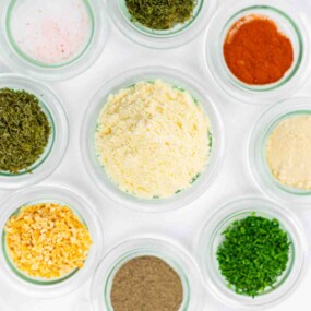 Nine small glass bowls containing various dried herbs, spices, and cheese powders are arranged on a white surface.