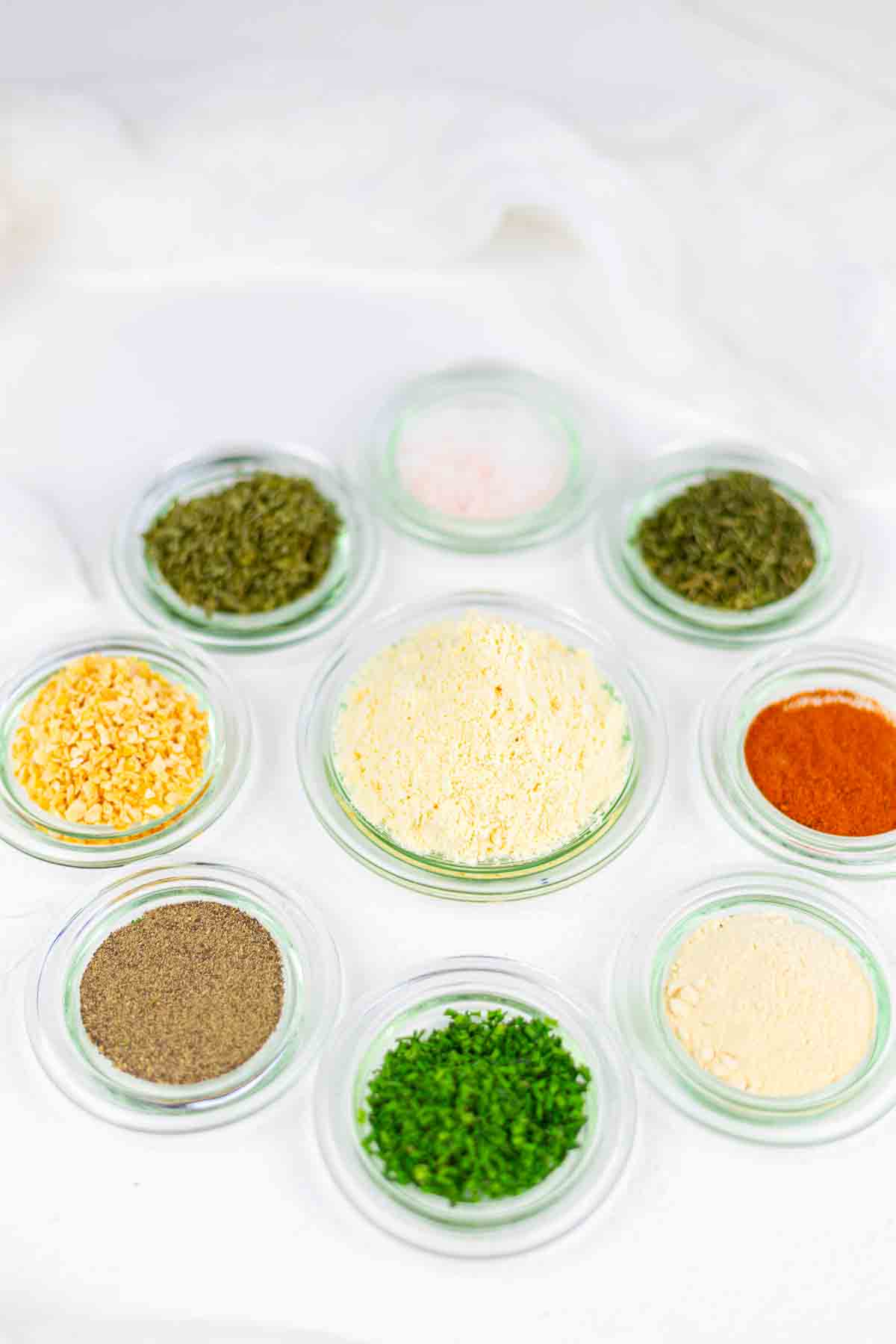 Nine small glass bowls with different dried spices and herbs, including paprika, black pepper, parsley, and garlic powder, arranged on a white surface.