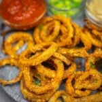 A plate of crispy breaded onion rings garnished with herbs, served with dips including salsa and a creamy sauce in small bowls.