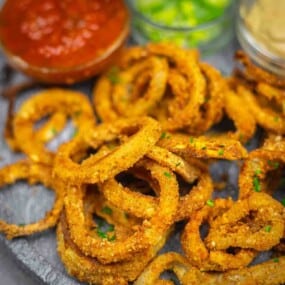 A plate of crispy breaded onion rings garnished with herbs, served with dips including salsa and a creamy sauce in small bowls.