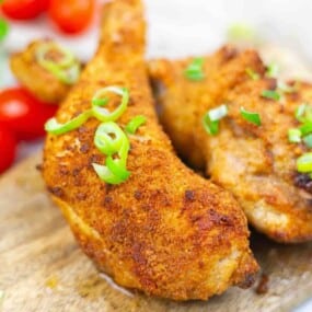 Close-up of crispy baked chicken legs garnished with sliced green onions, placed on a wooden surface with cherry tomatoes in the background.