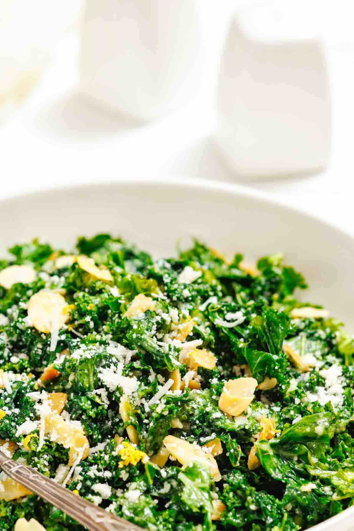 A bowl of kale salad topped with sliced almonds and grated cheese, with a fork resting inside the bowl.