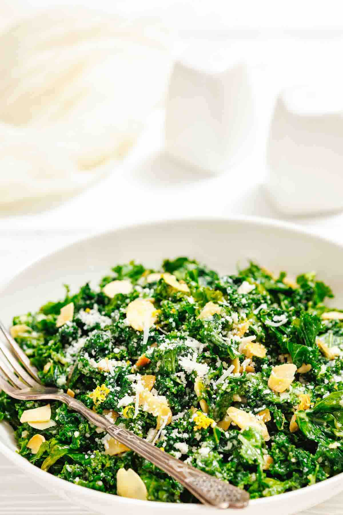 A white bowl filled with kale salad topped with shredded cheese, almond slices, and lemon zest, with a fork resting on the side.
