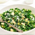 A white bowl filled with kale salad, topped with sliced almonds and grated cheese, with a fork resting on the side.