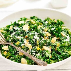 A white bowl filled with kale salad, topped with sliced almonds and grated cheese, with a fork resting on the side.