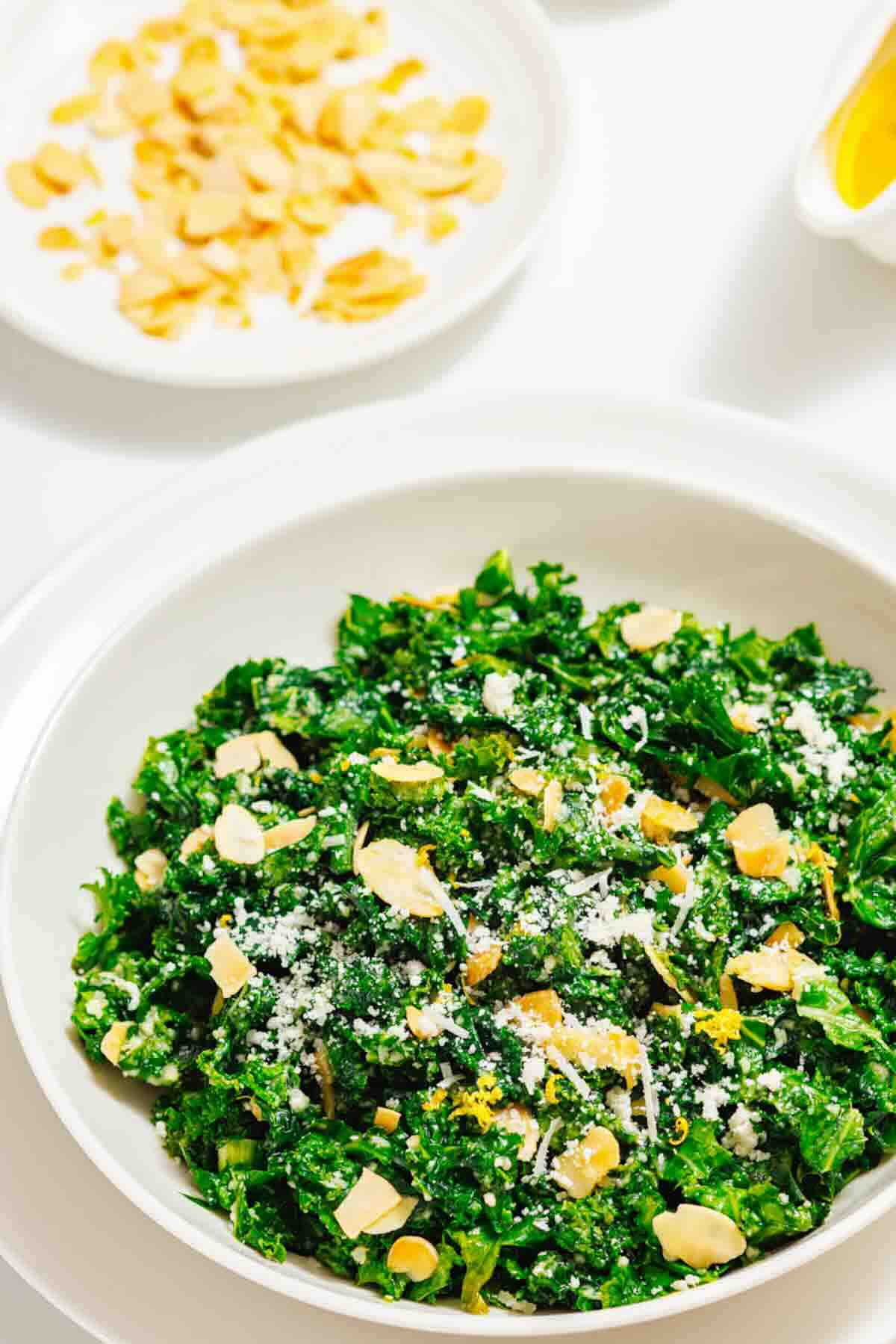 A white bowl filled with kale salad topped with sliced almonds and grated cheese, with a plate of almonds in the background.