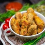 A bowl filled with golden-brown breaded and fried finger foods, garnished with herbs, with cherry tomatoes and a red dipping sauce in the background.