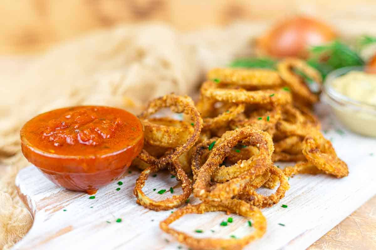 A pile of crispy fried onion rings is served on a white board with a small bowl of tomato-based dipping sauce and a yellow dipping sauce in the background.