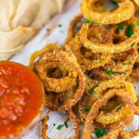 A pile of crispy fried onion rings garnished with chopped herbs sits next to a bowl of red dipping sauce on a wooden surface.