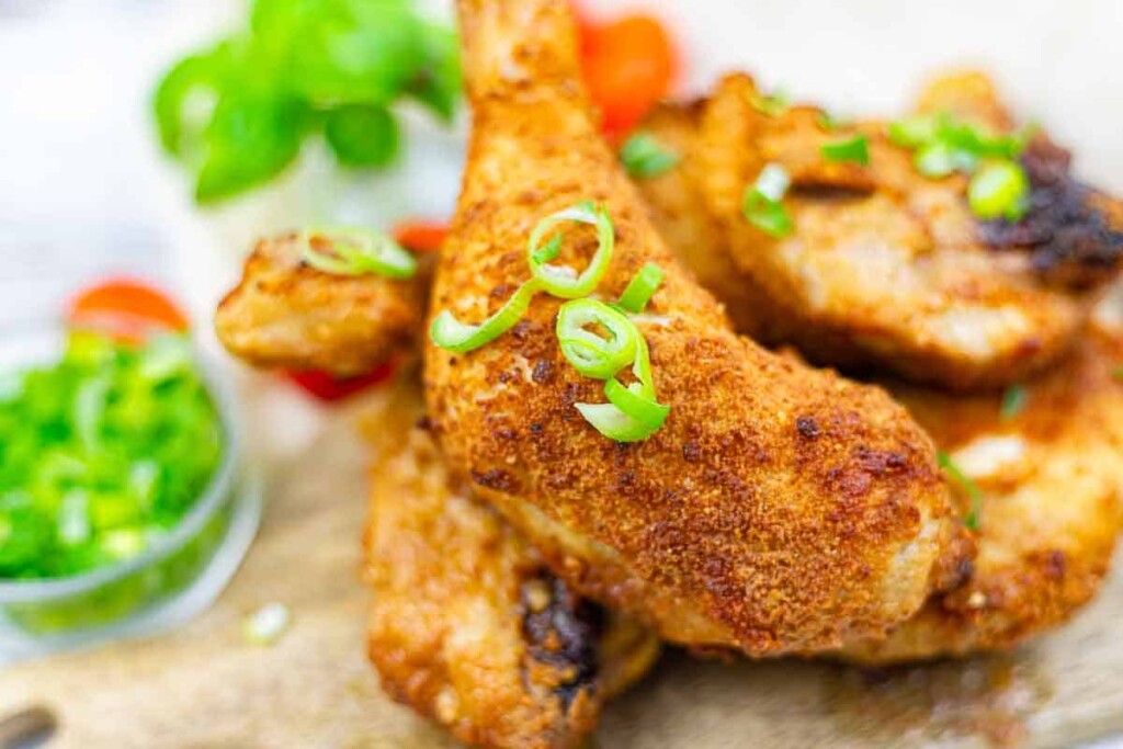 Close-up of crispy fried chicken pieces garnished with sliced green onions, with a side of chopped green onions and leafy greens in the background.