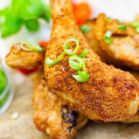 Close-up of crispy fried chicken pieces garnished with sliced green onions, with a side of chopped green onions and leafy greens in the background.