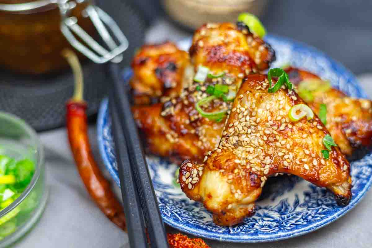 Grilled chicken wings topped with sesame seeds and chopped green onions, served on a blue patterned plate with chopsticks beside them.