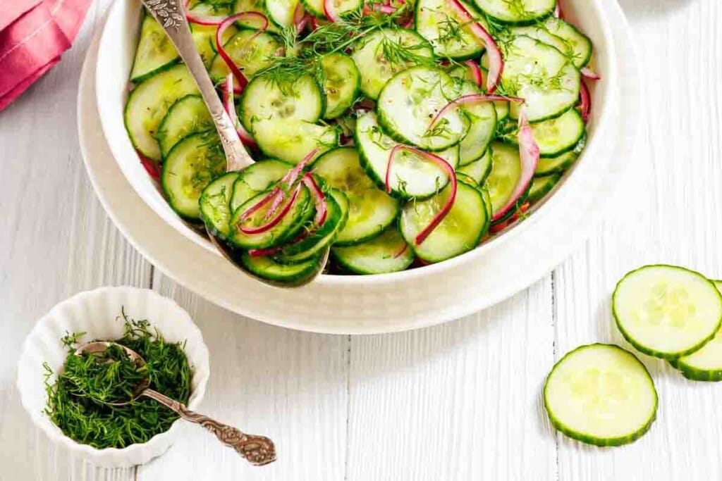 A bowl of cucumber salad with sliced red onions and fresh dill, next to a small dish of chopped dill and cucumber slices on a white wooden surface.