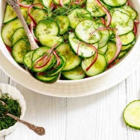A bowl of cucumber salad with sliced red onions and fresh dill, next to a small dish of chopped dill and cucumber slices on a white wooden surface.