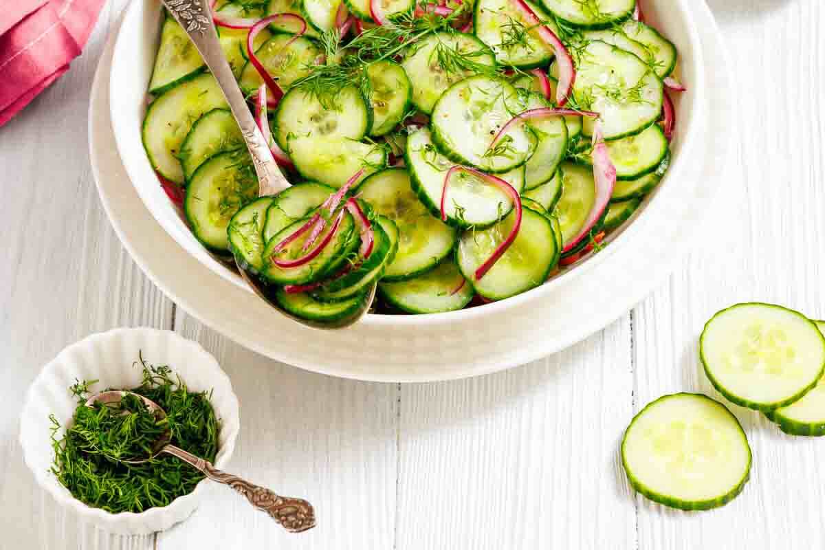 A bowl of cucumber salad with sliced red onions and fresh dill, next to a small dish of chopped dill and cucumber slices on a white wooden surface.