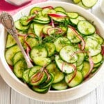 A white bowl filled with sliced cucumber, red onion, and fresh dill salad, with a silver spoon resting inside.