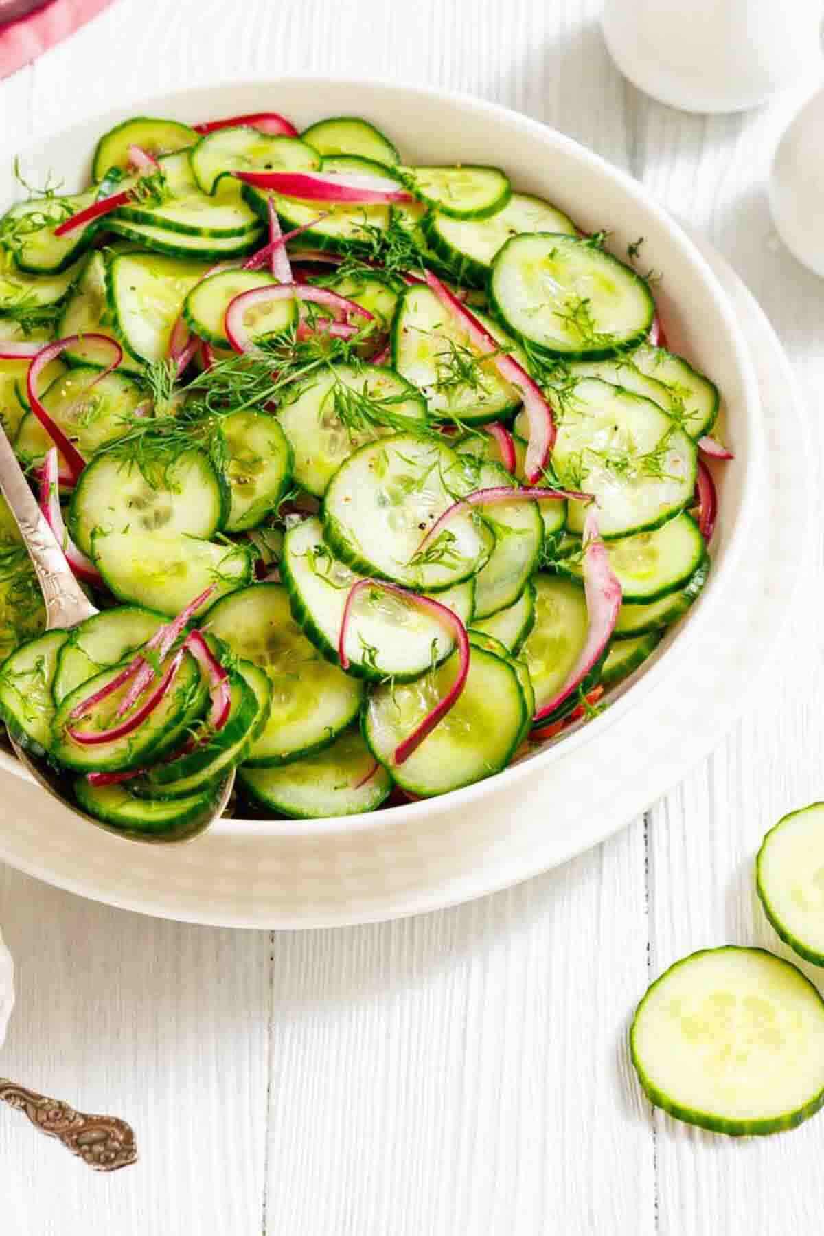 A bowl of cucumber salad with sliced red onions and fresh dill, served on a light wooden surface.