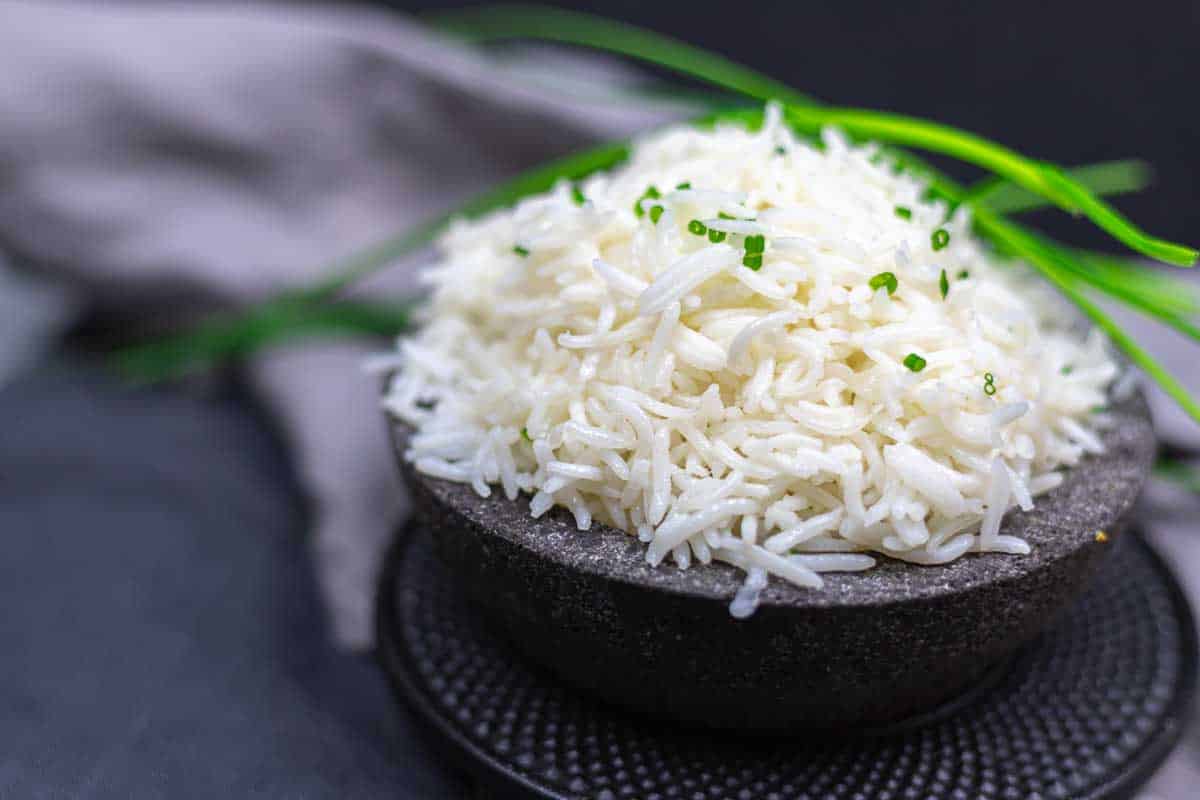 A bowl of cooked white rice garnished with chopped green herbs, placed on a dark plate with a gray cloth and green herbs in the background.