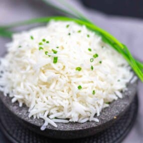 A mound of cooked white basmati rice garnished with chopped green herbs, served on a dark round plate with whole green herbs placed on top.