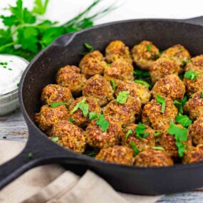 A cast iron skillet filled with cooked meatballs garnished with parsley, with a bowl of white dipping sauce and fresh herbs in the background.