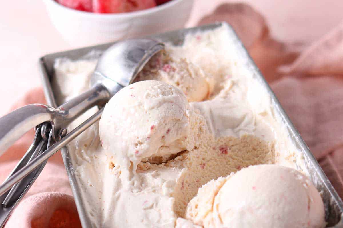 Rectangular metal container filled with scoops of creamy ice cream and a metal ice cream scoop, with a bowl of fruit in the background.