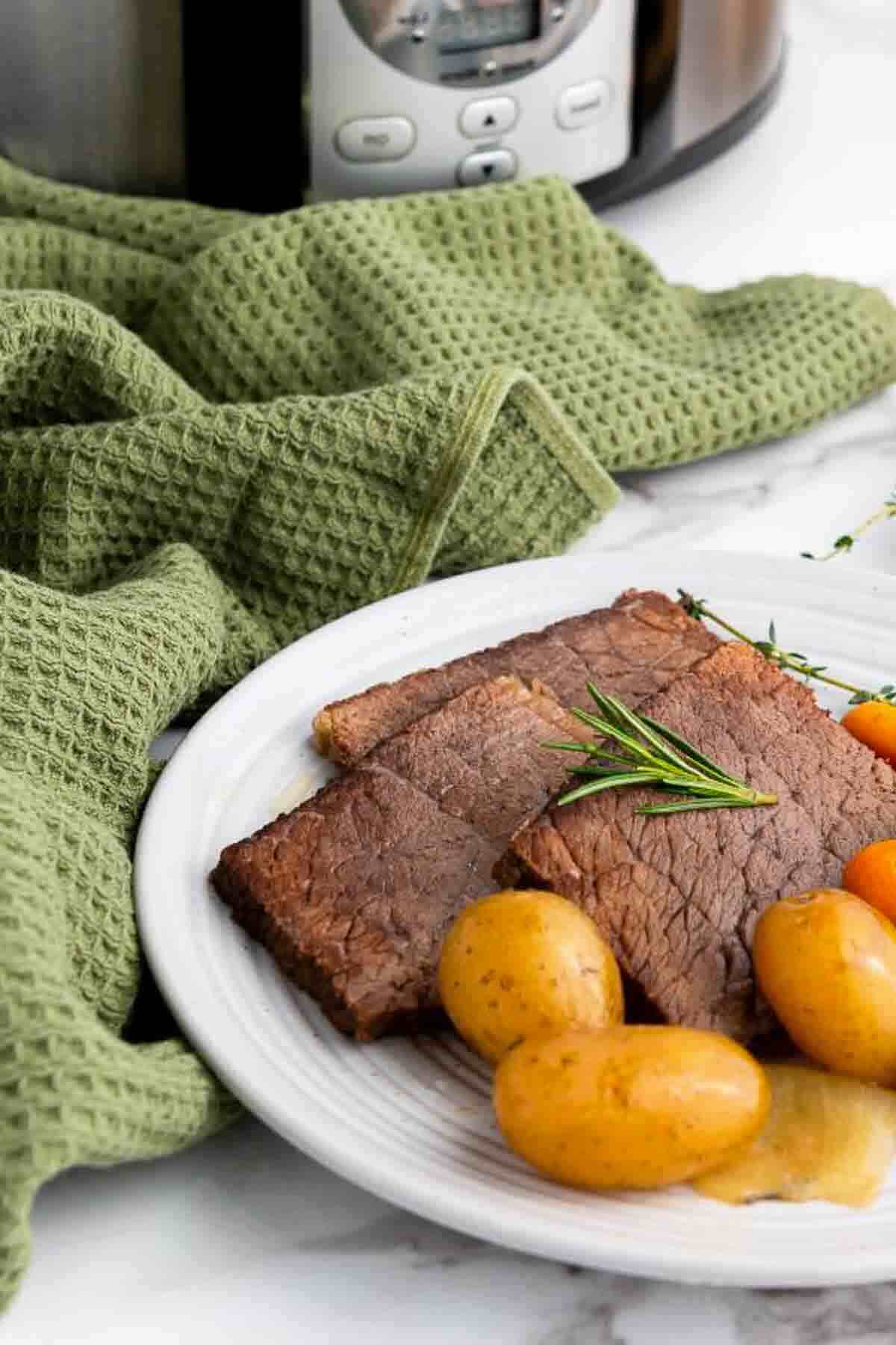 A plate with slices of pot roast, baby potatoes, and carrots, garnished with rosemary, sits in front of a green towel and a slow cooker.