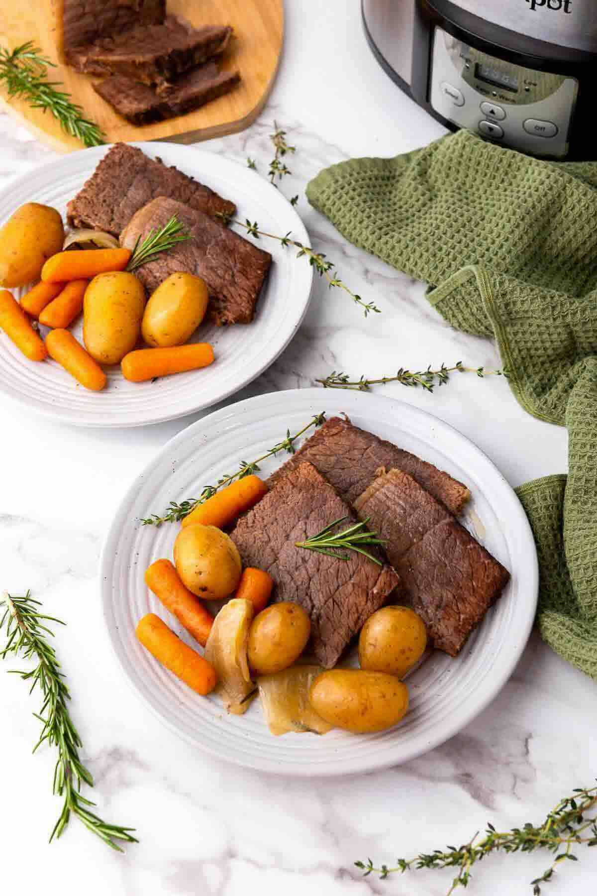 Two white plates with sliced pot roast, whole baby potatoes, carrots, and onion, garnished with rosemary, placed on a marble countertop near a green towel and a slow cooker.