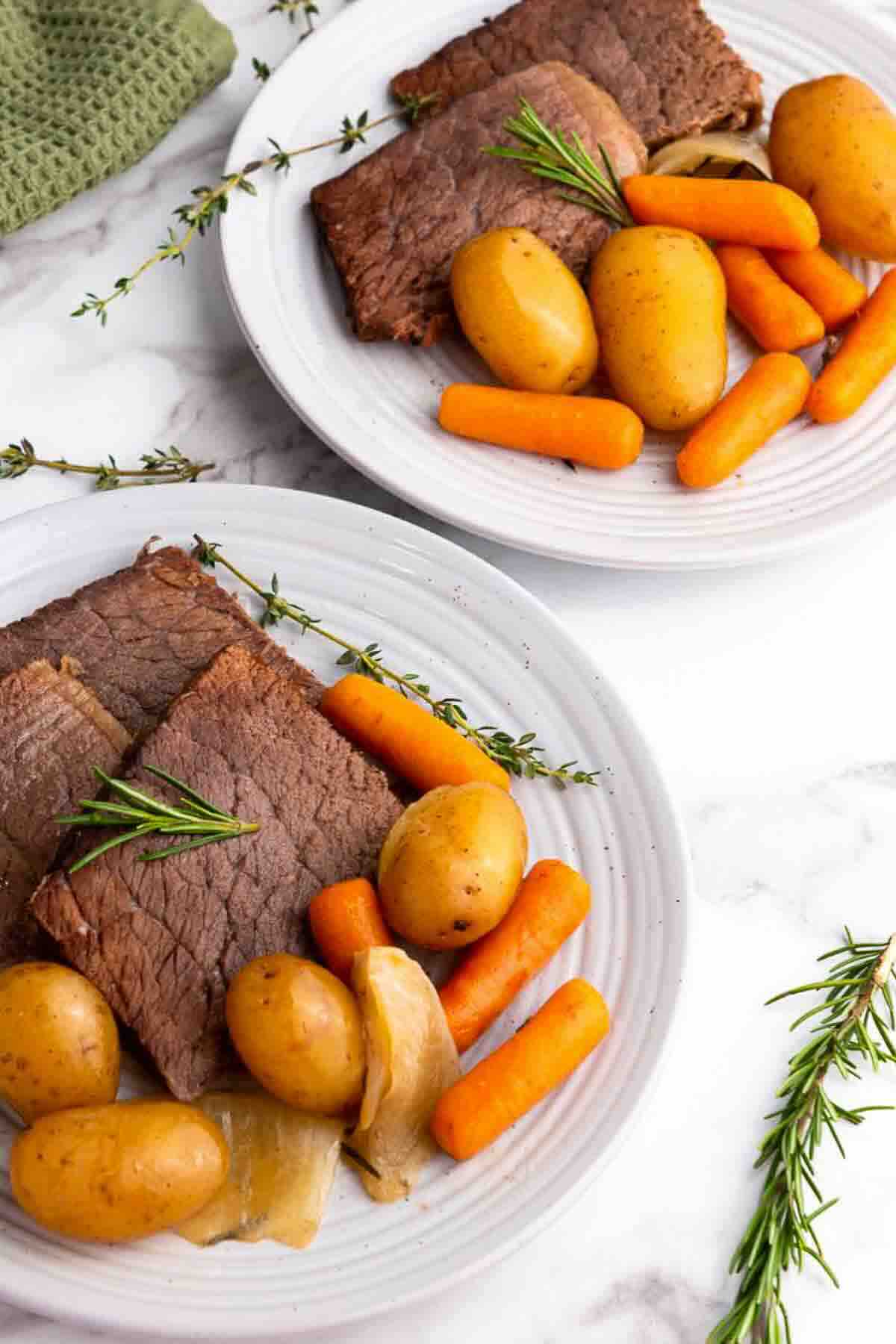 Two white plates with slices of roast beef, baby potatoes, carrots, and onions, garnished with fresh rosemary and thyme on a white surface.