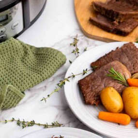 A plate with sliced pot roast, potatoes, and carrots sits on a marble surface next to a slow cooker, a green towel, and fresh herbs.
