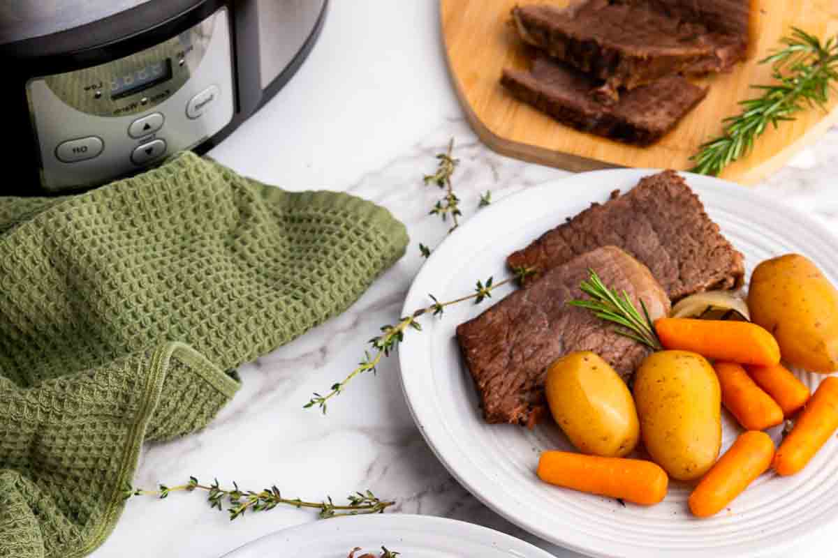 A plate with sliced pot roast, potatoes, and carrots sits on a marble surface next to a slow cooker, a green towel, and fresh herbs.