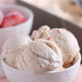 Three scoops of strawberry ice cream in a white bowl, with a bowl of frozen strawberries and an ice cream container in the background.