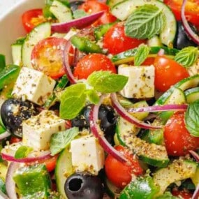 A bowl of salad with tomatoes, cucumbers, olives, red onion, feta cheese cubes, and fresh mint leaves, seasoned with herbs.