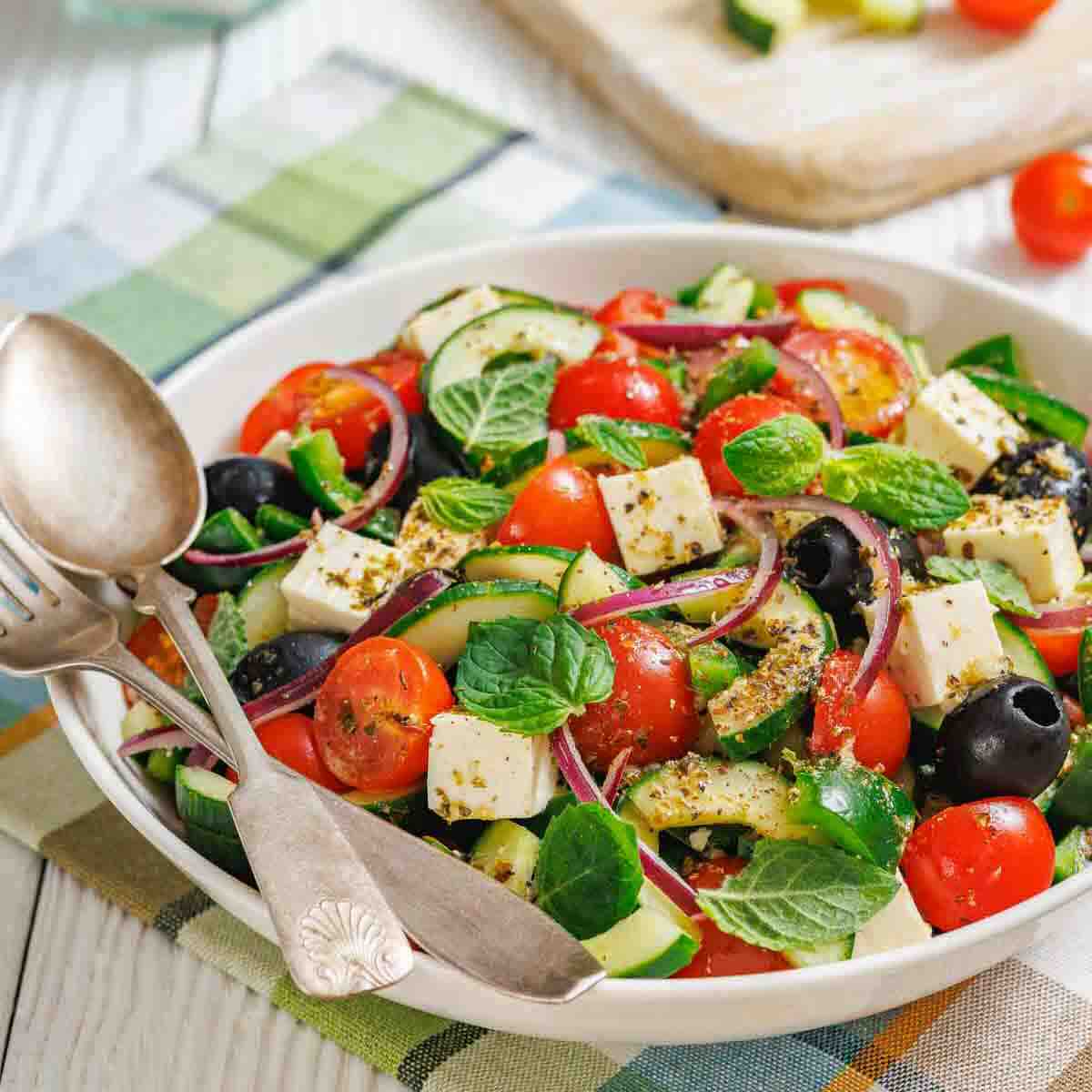 A bowl of Greek salad with cherry tomatoes, cucumbers, feta cheese, black olives, red onions, and fresh mint leaves, with serving utensils resting on the side.