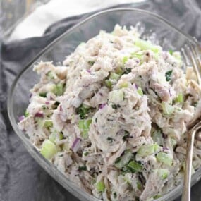 A glass bowl filled with chicken salad mixed with chopped celery, red onion, and herbs, with a fork on the side.