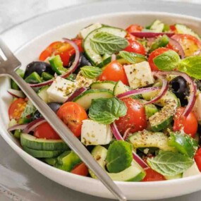 A bowl of Greek salad with cherry tomatoes, cucumbers, olives, feta cheese, red onions, and fresh mint leaves, with a fork resting on the plate.