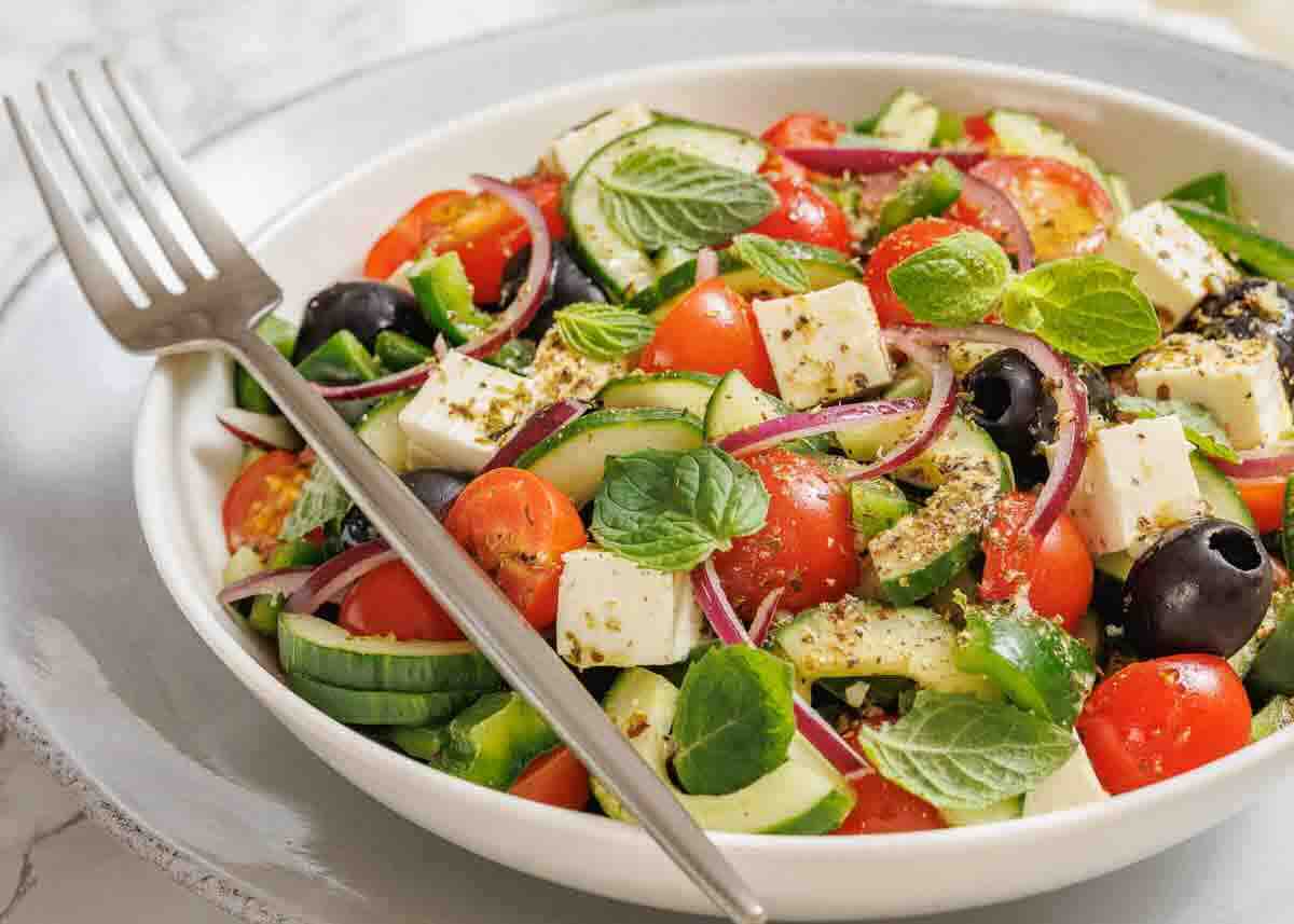 A bowl of Greek salad with cherry tomatoes, cucumbers, olives, feta cheese, red onions, and fresh mint leaves, with a fork resting on the plate.