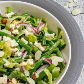 A bowl of salad with green beans, lettuce, sliced red onions, crumbled feta cheese, sliced almonds, and chopped walnuts.