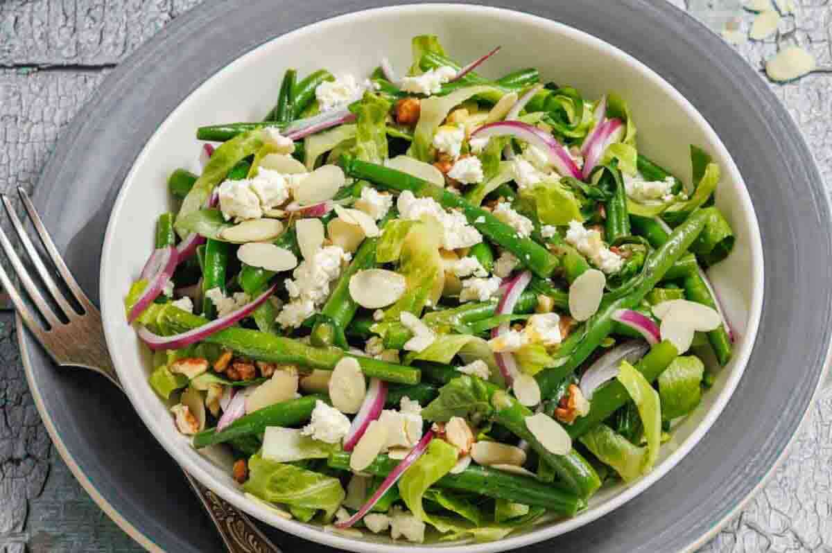 A bowl of green bean salad with sliced red onions, slivered almonds, goat cheese, lettuce, and walnuts, placed on a gray plate with a fork beside it.