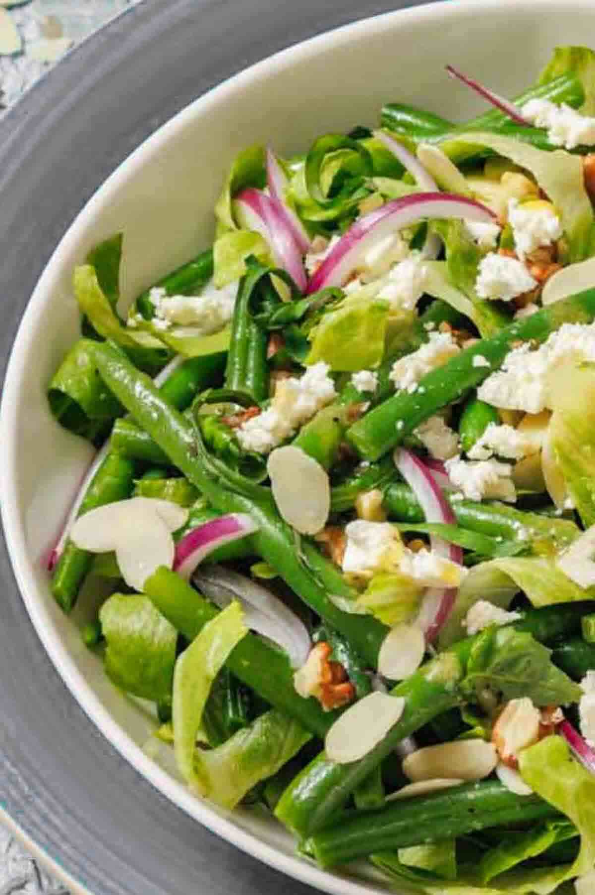 A bowl of green bean salad with sliced red onions, crumbled feta cheese, slivered almonds, and mixed greens.