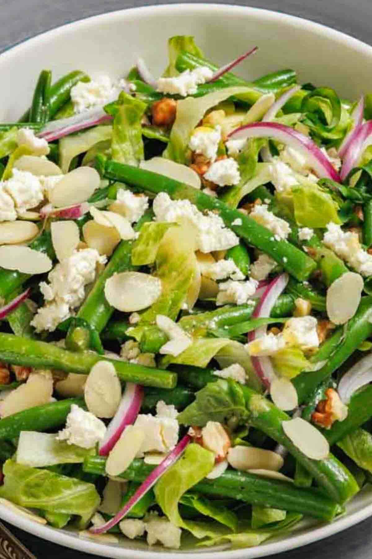 A bowl of salad containing green beans, lettuce, feta cheese, red onion slices, almonds, and walnuts.