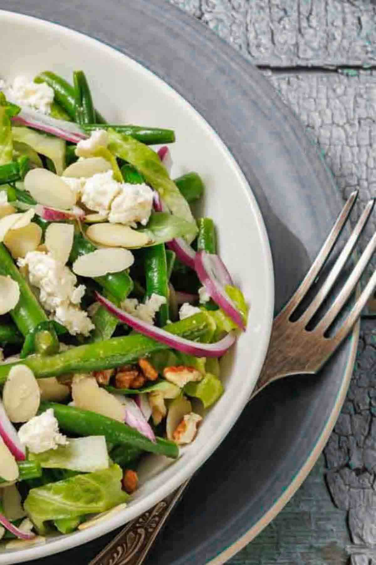 A close-up of a salad with green beans, red onion, sliced almonds, crumbled cheese, and lettuce in a white bowl on a plate with a fork.