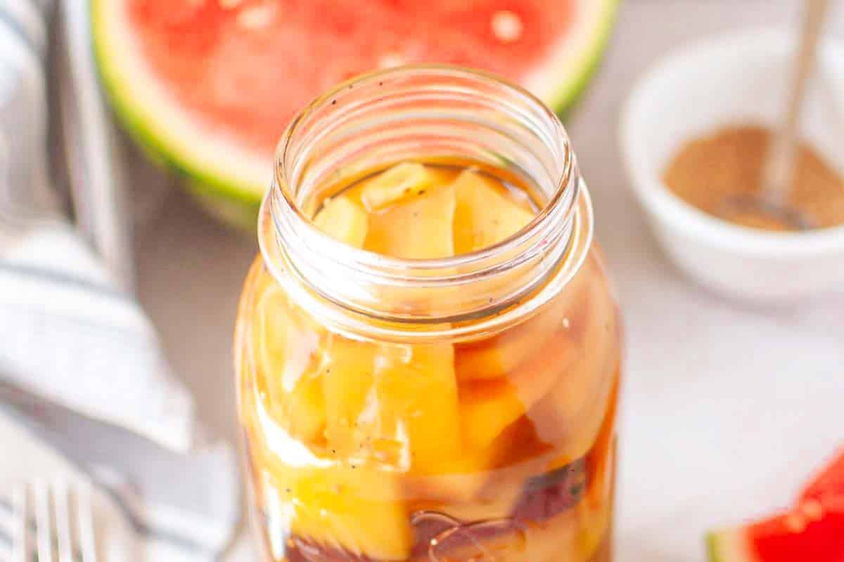 A glass jar filled with pickled watermelon rind sits on a light surface; a watermelon slice and a small bowl of brown seasoning are in the background.