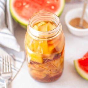 A glass jar filled with pickled watermelon rind sits on a light surface with slices of fresh watermelon and a bowl of spices in the background.
