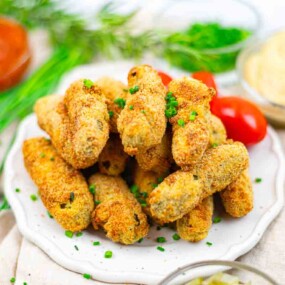 A plate of golden-brown, breaded croquettes garnished with chopped chives, with cherry tomatoes and dips in the background.