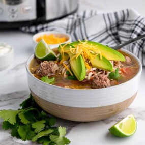 A bowl of soup with beef, avocado slices, shredded cheese, and cilantro, with a lime wedge and fresh cilantro beside it; slow cooker, sour cream, and shredded cheese in the background.