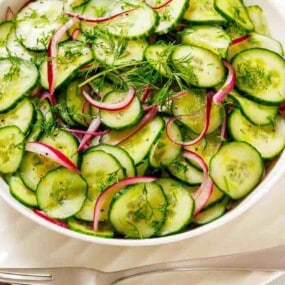 A bowl of sliced cucumber salad with red onions and fresh dill sits on a white plate beside a fork and spoon.