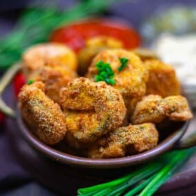 A bowl of breaded and fried mushrooms garnished with chopped herbs, with dips and fresh vegetables in the background.