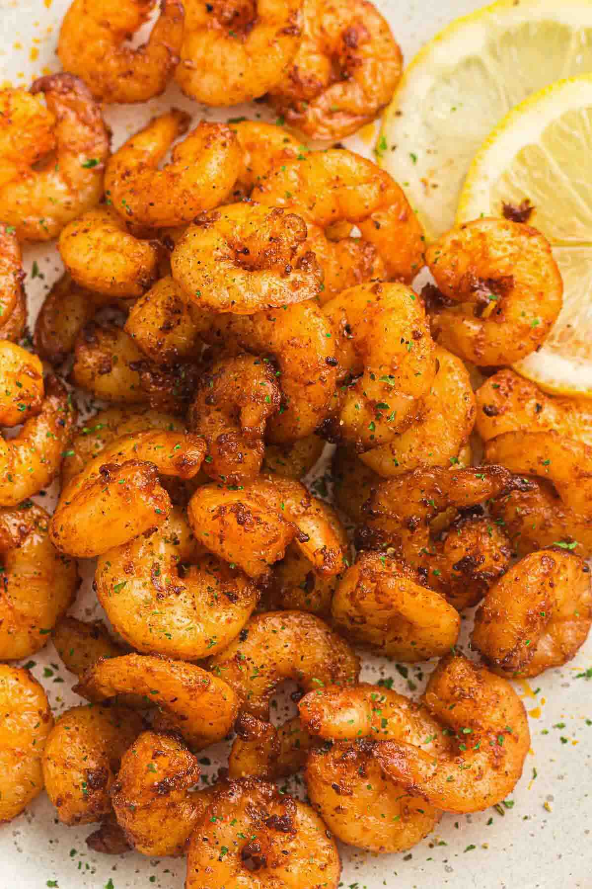 Close-up of seasoned, cooked shrimp garnished with herbs, served with thin lemon slices on a light-colored plate.